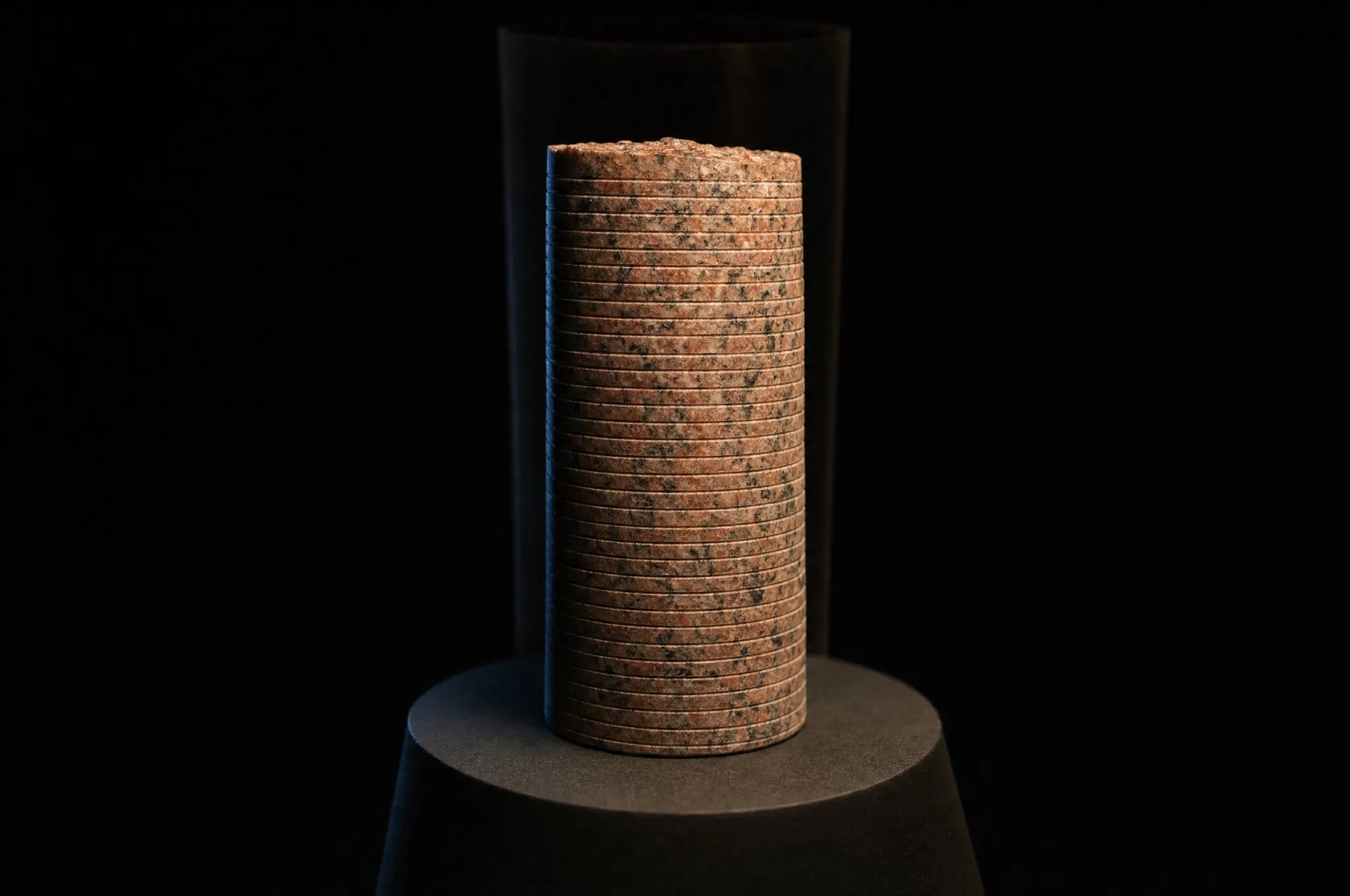 A museum-lit close study of a tapered cylinder of rose-red Aswan granite standing on a dark pedestal against blackness: a hand-sized ancient drill core, its speckled surface crossed by a fine shallow groove winding around the stone with about a millimetre and a half between traces, a warm key light raking across the crystals from the upper right, a faint translucent ghost of a copper tube drill enclosing the core, and darkness beyond the pedestal edge.