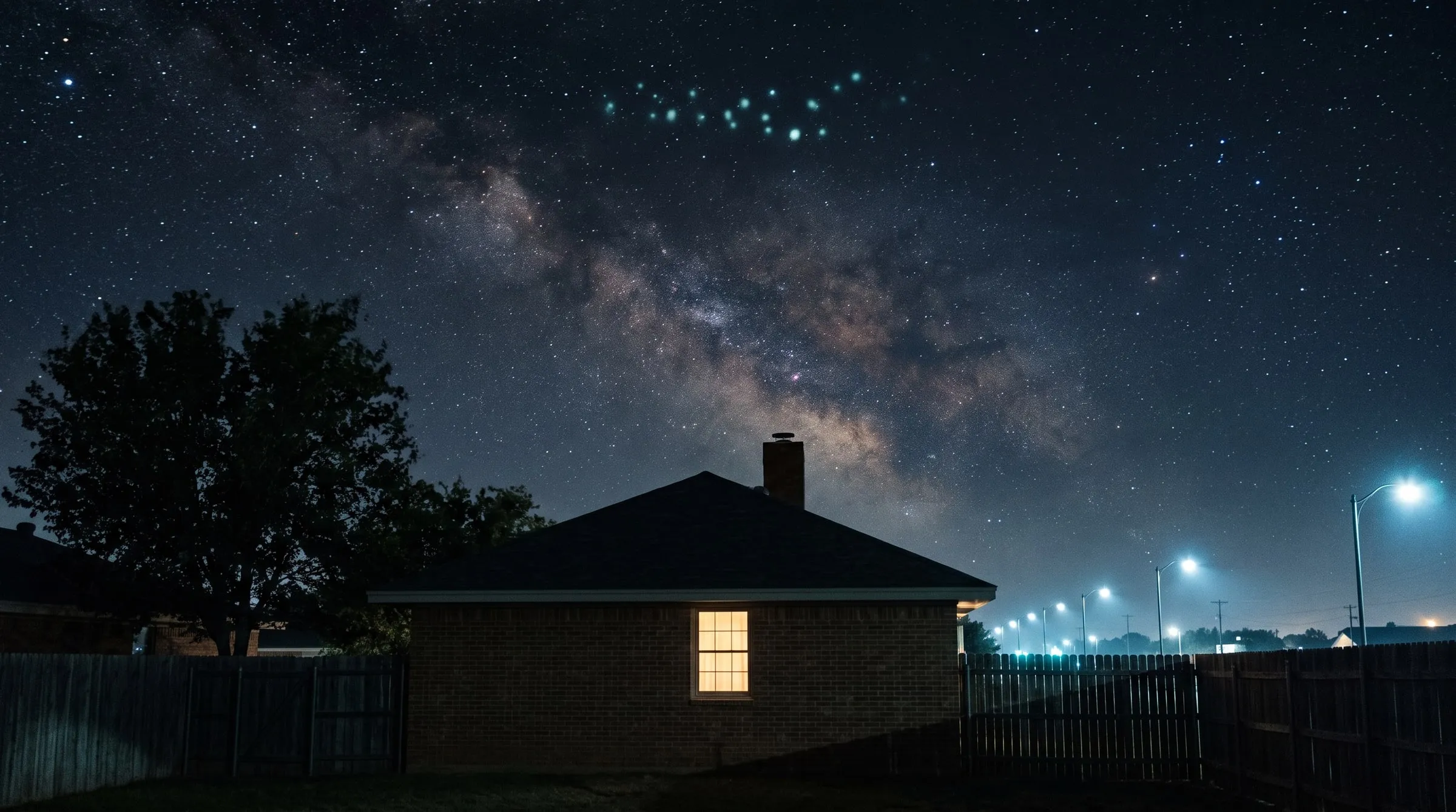 A suburban backyard on a clear night seen from the lawn, looking up past the hip roof and brick chimney of a low brick house with one warm lit window: a wide sky full of stars with the Milky Way rising from behind the roof toward the upper left, and high above it a small loose chevron of about twenty soft bluish-green glowing points, its point downward toward the house; a wooden fence closes the yard on both sides, a leafy tree stands at the left, and beyond the right-hand fence a line of tall street lamps on curved arms gives a pale bluish-white light with a haze of city glow low on that horizon; no Moon, no people, no text.
