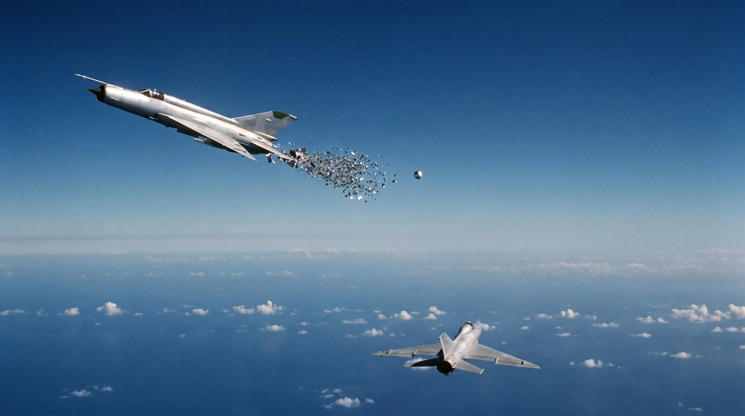 High over a blue tropical sea dotted with small clouds, a silver MiG-21 crosses the upper left of the frame, its rear fuselage dissolving into a comet-tail of thousands of glittering metal fragments streaming down and back through the air, with no fire and no smoke anywhere; a small bright metallic sphere hangs in the sky to the right of the trail; in the lower right an identical wingman jet flies on, seen from behind and above; hard daylight, no people visible, no legible text.