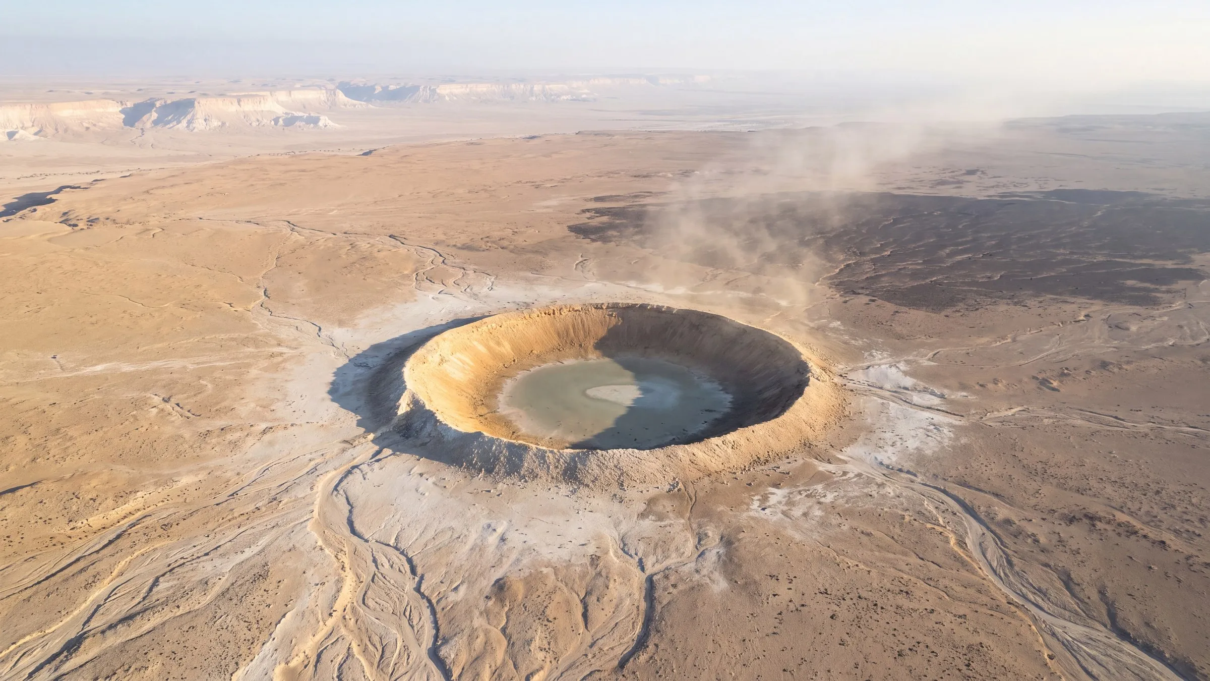 High-oblique aerial view over a pale limestone plateau in the central Sinai at early morning: one vast fresh crater cut into the ochre gravel plain, its raised rim of thrown-out rubble catching hard sunlight from the right, the sunlit inner wall bright and the far wall in deep shadow, the floor a smooth pale grey-green glassy sheet with a low mound at its centre; the ground around the crater bleached paler than the plain, dry braided wadis running across the foreground; to the upper right a broad darker plain of flint gravel with a thin veil of dust drifting away from the crater toward the hazy horizon; white limestone escarpments in the far distance; no people, no text.