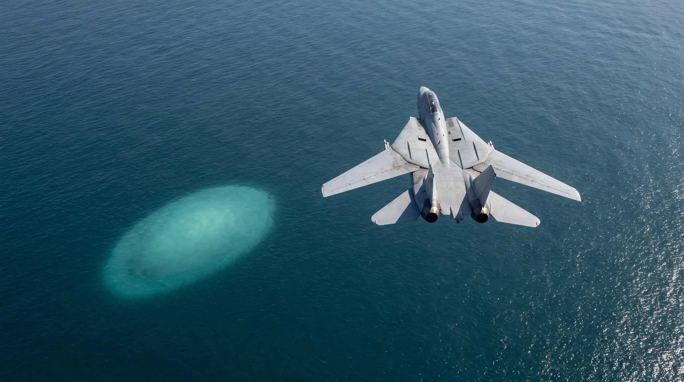 Late morning over the open Pacific, seen from high above: a grey F-14D Tomcat with its wings part-swept flies away from the viewer toward the top of the picture, right of centre, in a gentle bank, the crew's shapes just visible inside the canopy; off its left wing and lower in the frame, a pale milky-teal egg-shaped patch lies just beneath the calm blue-green surface, brightest toward its centre and fading into the water at its edges, the sea's fine ripples continuing across it; the water darkens toward the left and carries a broad field of sun glitter toward the right; no horizon, no coast, no ships, no one outside the aircraft, no text.