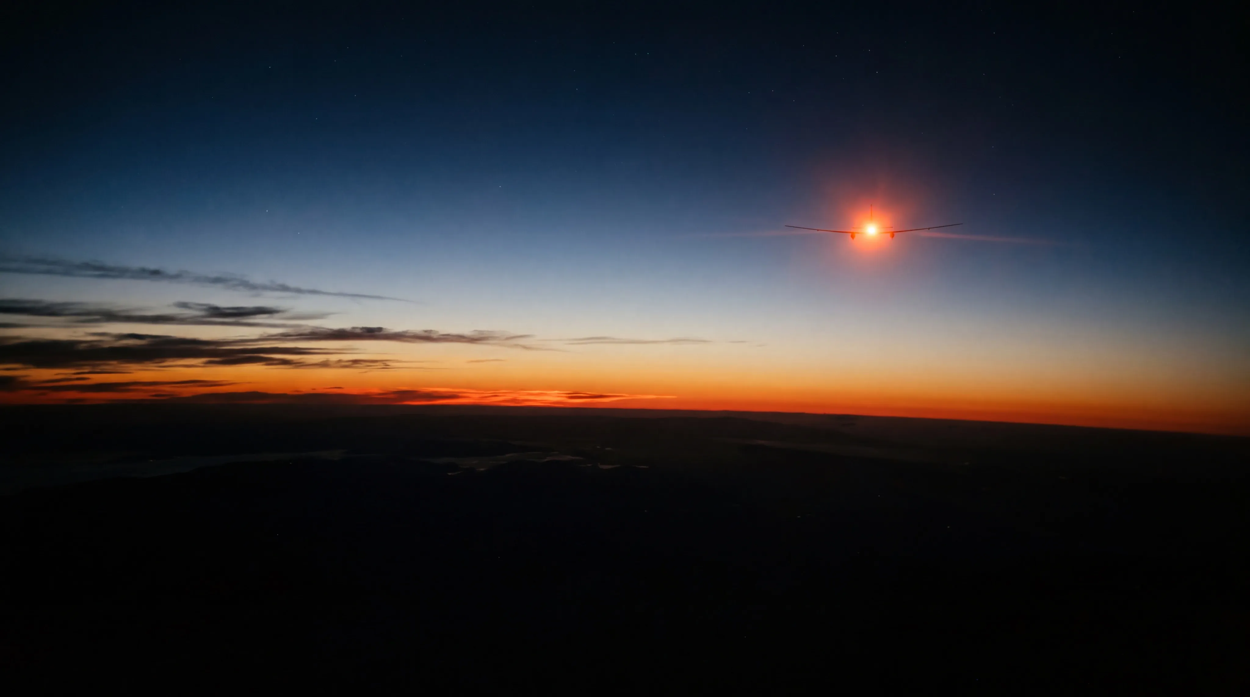 A wide view from high altitude at deep dusk: the Earth's horizon is a long gently curved dark edge across the lower third of the frame with absolutely black unlit land below it, and above it a narrow band of burnt orange and ember red fades upward through amber into slate blue and then into a deep indigo sky flecked with a few faint stars; high in the upper right, a single small brilliant point of fiery orange light burns against the darkening blue, far brighter than any star, with the faint slender silhouette of very long thin straight wings just resolvable around it and a small warm halo of glare, and nothing else in the sky is lit.